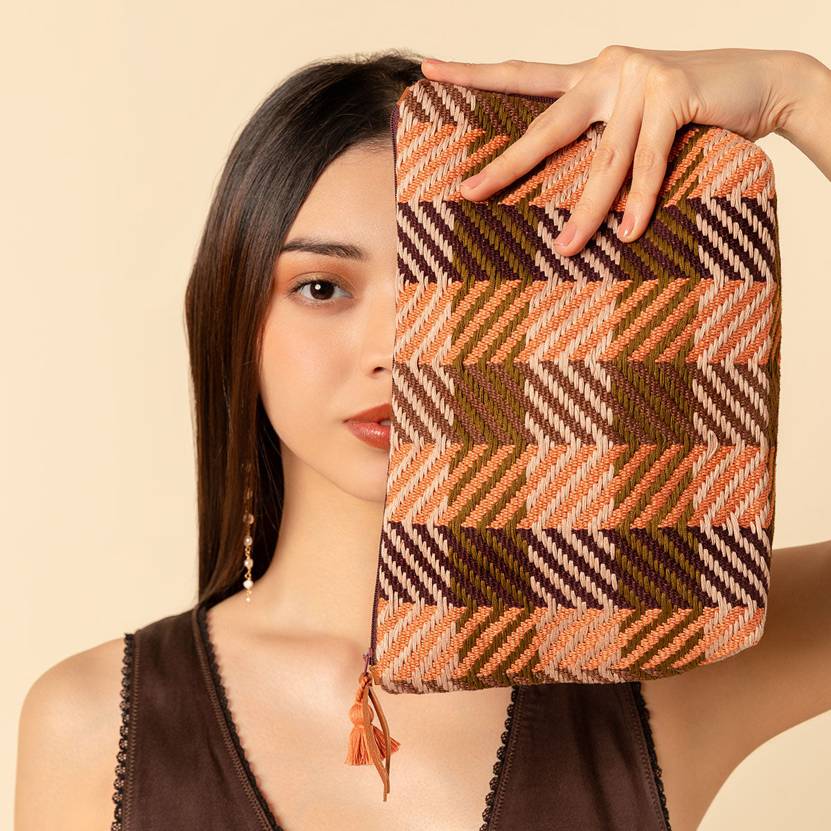 Hand woven artisan Cristina Cosmetic Pouch in Moss Weave pattern. A model holds the Cristina pouch vertically in front of her head, showing half of her face. The Cristina has an orange and olive green plaid, an orange tassel, and a leather zipper pull.