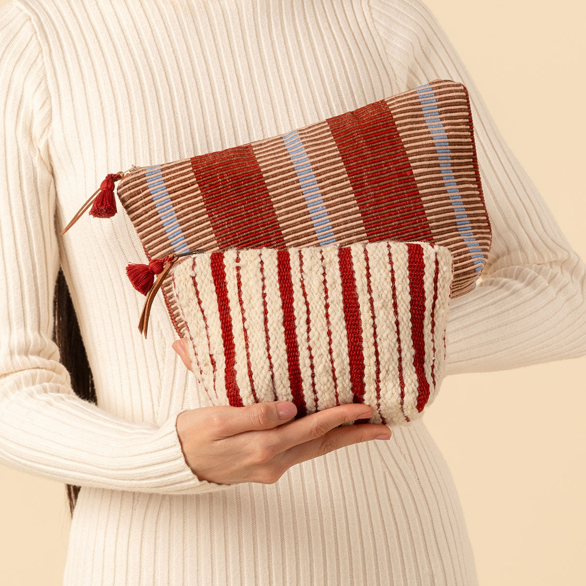 Hand woven artisan Cristina Cosmetic Pouch in Chestnut and Mini Cristina Pouch in Cherry Red. A model holds both bags. The Mini Cristina has vertical red and beige stripes for the Cherry Red color. The Cristina has Chestnut colors, thick burgundy and tan vertical stripes.