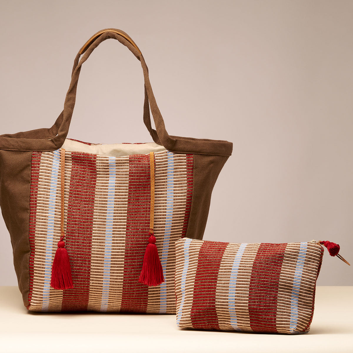 Artisan hand woven Cristina Cosmetic Pouch and Rose Tote in Chestnut color are placed together. The Rose tote has red tassels attached to leather cords. The Cristina Pouch has a mini red tassel and a leather zipper pull. The Chestnut color has thick vertical stripes in burgundy and tan.