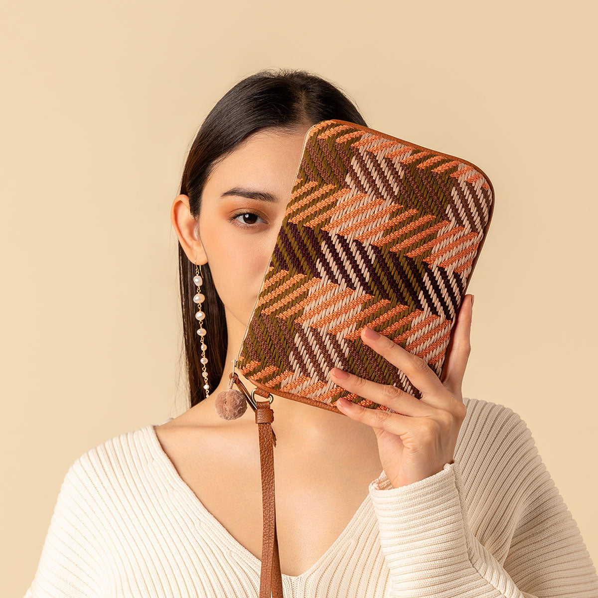 Hand woven artisan Mini Lily Wristlet in Moss Weave. A model holds up the Mini Lily diagonally over her face, exposing one eye. The Moss Weave has an orange and olive green plaid pattern. It has a leather handle and a pink pompom.