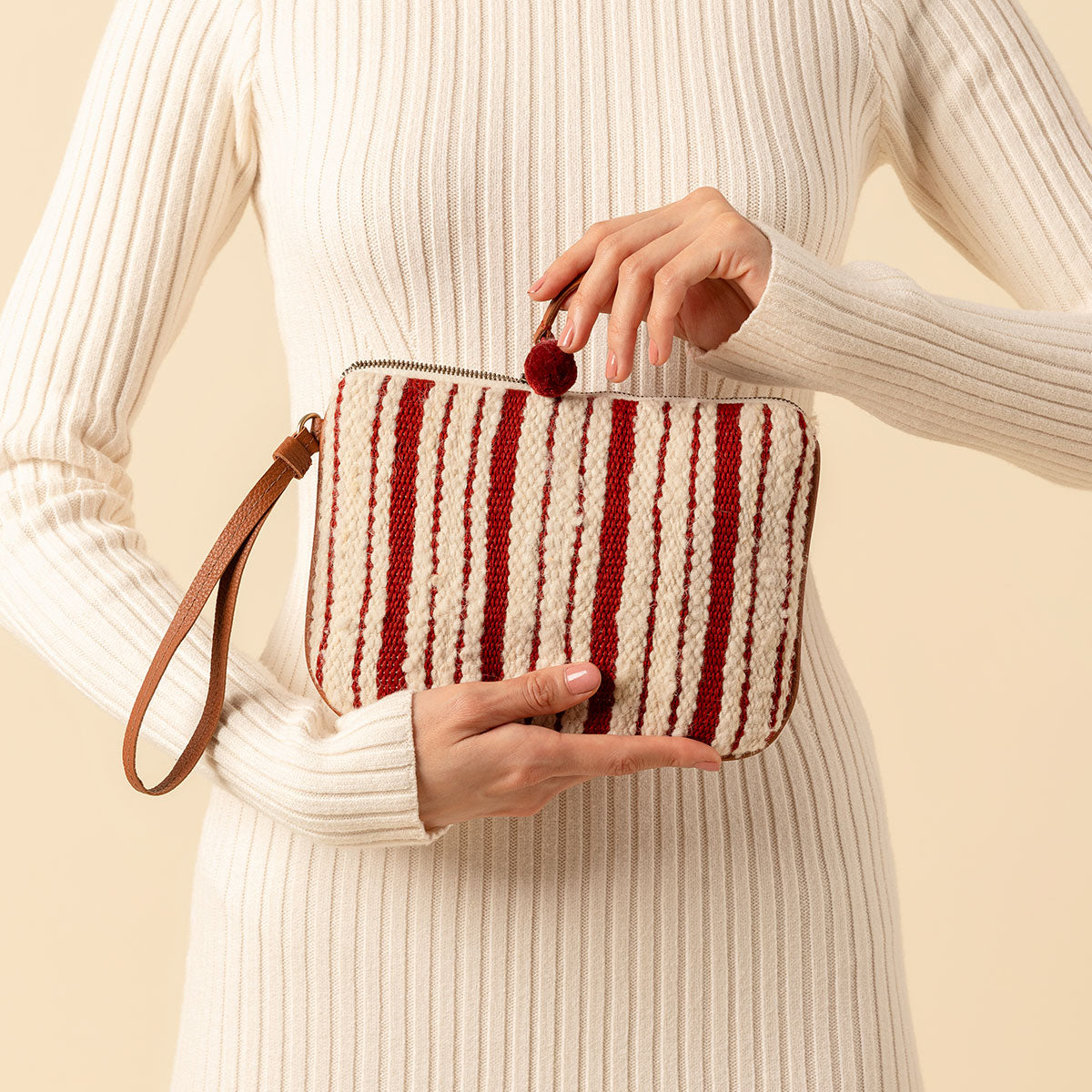 Artisan hand woven Mini Lily Wristlet in Cherry Red pattern. It has vertical red and beige stripes, a leather hand strap and a red pompom attached to the zipper. A model holds the Mini Lily Wristlet in her hand and opens the pouch with the zipper.