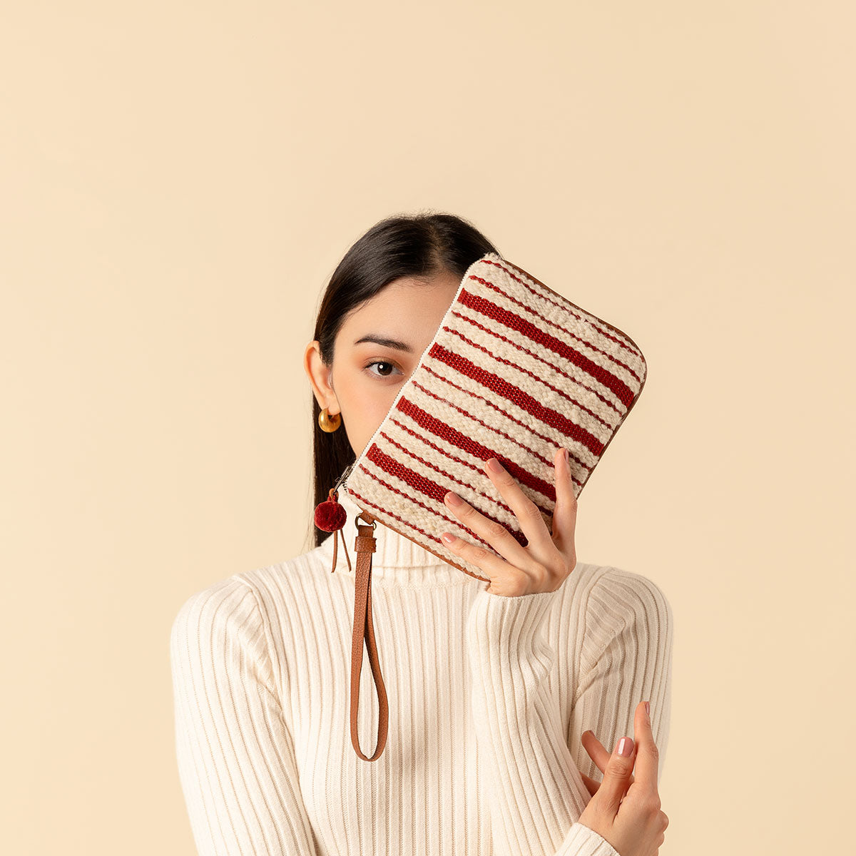 Artisan hand woven Mini Lily Wristlet in Cherry Red pattern. It has vertical beige and red stripes, a leather strap, and a red pompom. A model tilts the Mini Lily Wristlet over her face, showing one eye to the camera.