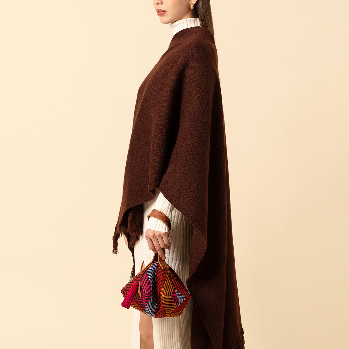 A model holds the Dulce Wristlet in Winter Berry pattern. The model is turned to the side profile and wears a brown shawl and a white sweater dress. The background is a cream yellow color. The Dulce Wristlet has a leather handle and a red tassel.