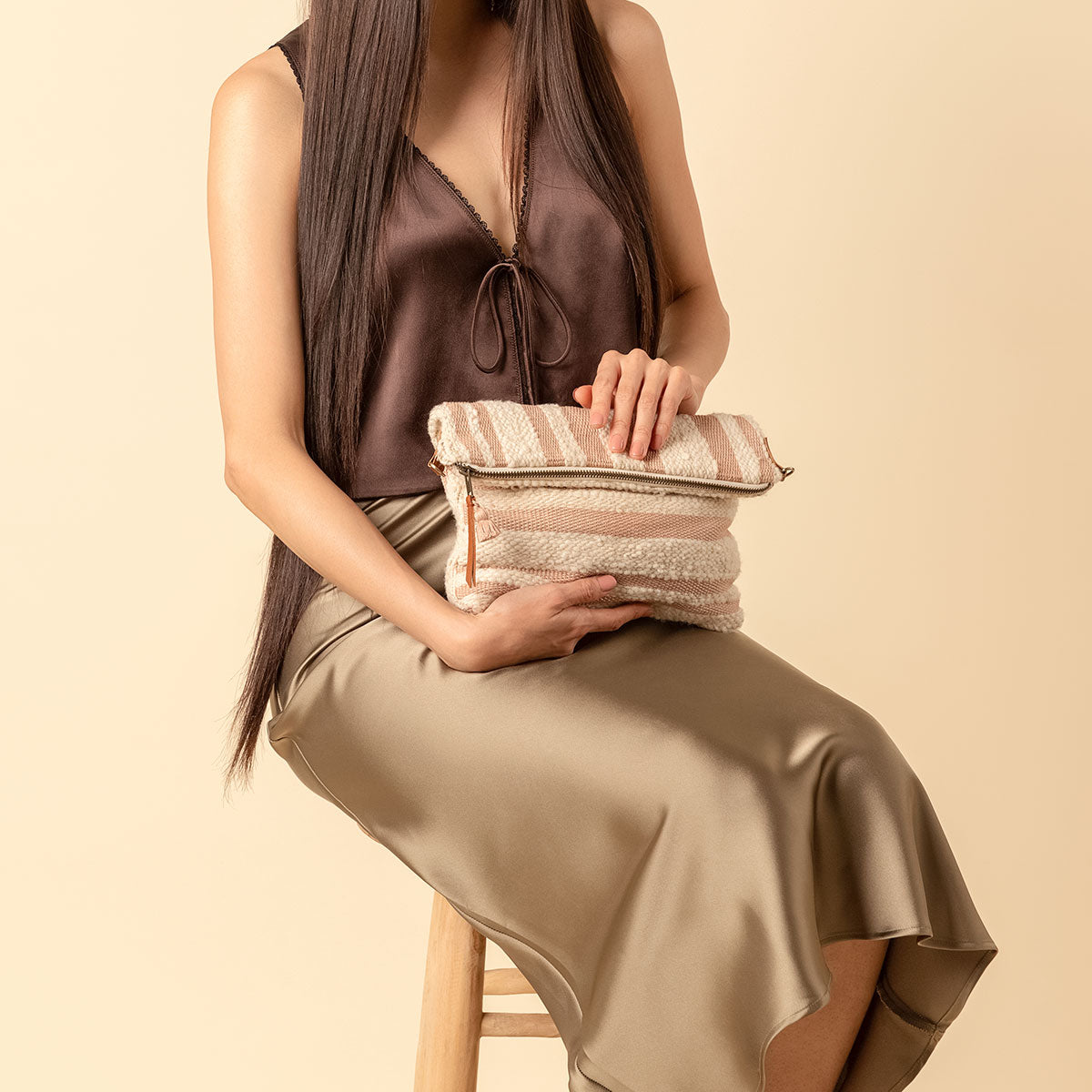 Hand woven artisan Florentina Clutch-to-hobo bag in Snow. A model sits on a stool and holds the folded Florentina in her lap at an angle. The bag has vertical and horizontal pink and beige stripes.