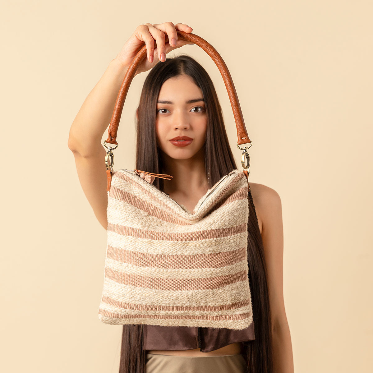 Hand woven artisan Florentina Clutch-to-Hobo bag in Snow. A model holds the bag with the leather handle framing her head. The unfolded Florentina has horizontal pale pink and beige stripes. It has a leather zipper pull and pale pink tassel.