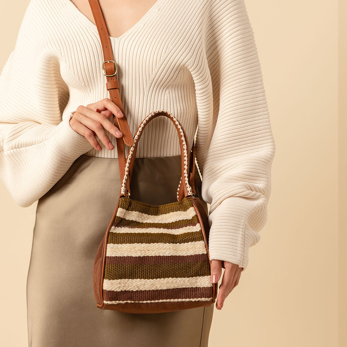 Hand woven artisan Flora Petite Tote in Forest shown at an angle. The Forest pattern has horizontal green and beige stripes. The leather handles have embroidered white detailing. A model wears the crossbody over her torso, an adjustable leather strap.