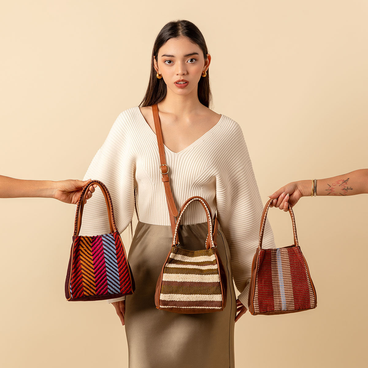 Hand woven artisan Flora Petite totes in three patterns: Winter Berry, Forest, and Chestnut. A model wears the Flora Petite in Forest over her torso. A hand holds the Winter Berry at the left and another holds the Chestnut at the right.