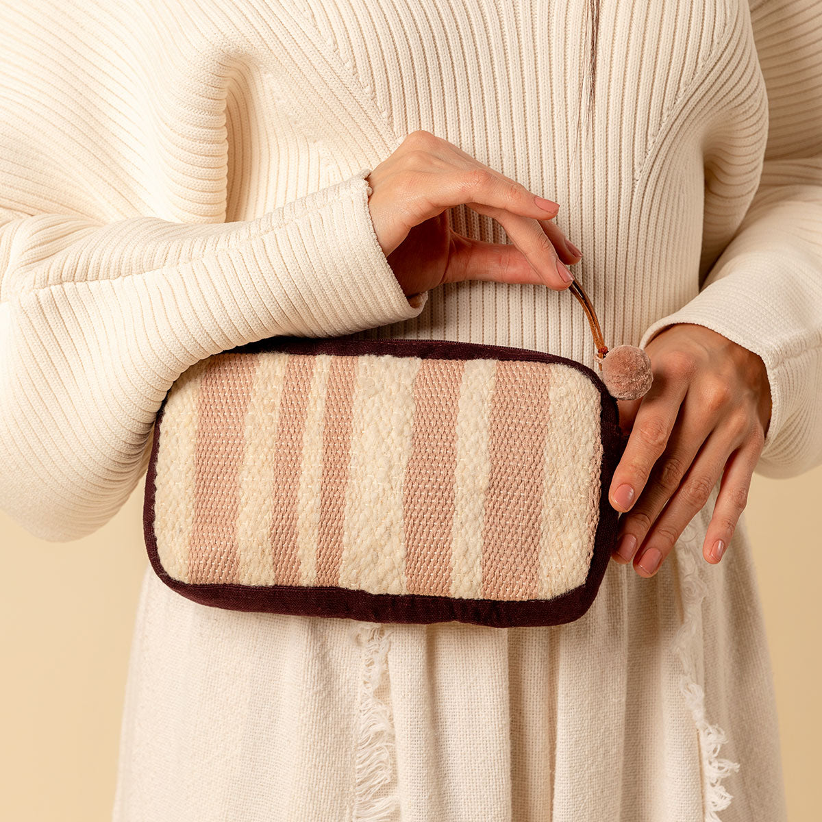 Hand woven artisan Felipa Bag in Snow pattern. A model holds the bag. It has vertical pale pink and beige stripes, a leather zipper and a pale pink pompom.