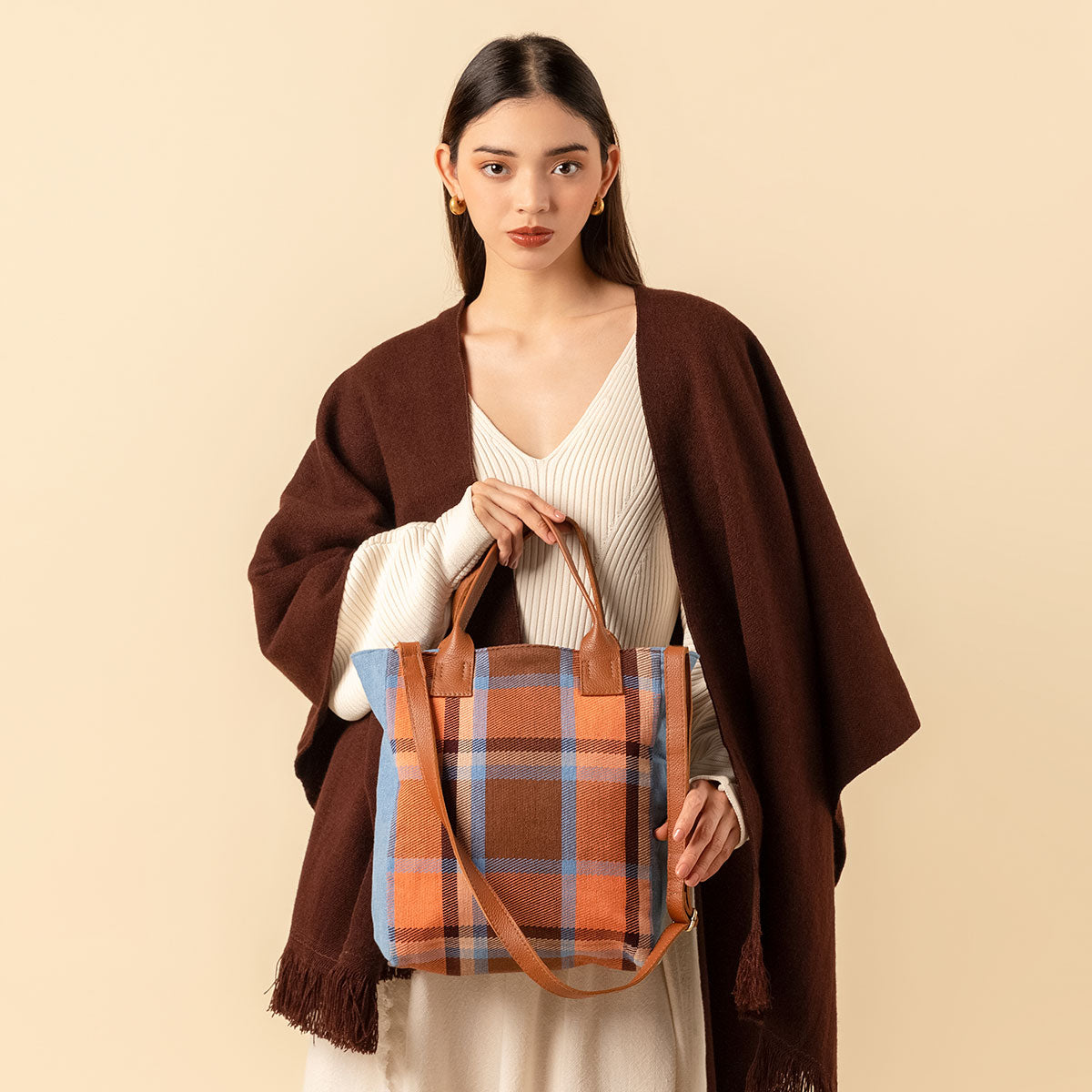 A model holds the handles of the hand woven artisan Ana tote in Brickstone pattern and looks at the camera. A leather adjustable strap hangs diagonally over the front. It has leather handles. The Brickstone pattern is a brown, orange, and sky blue plaid.