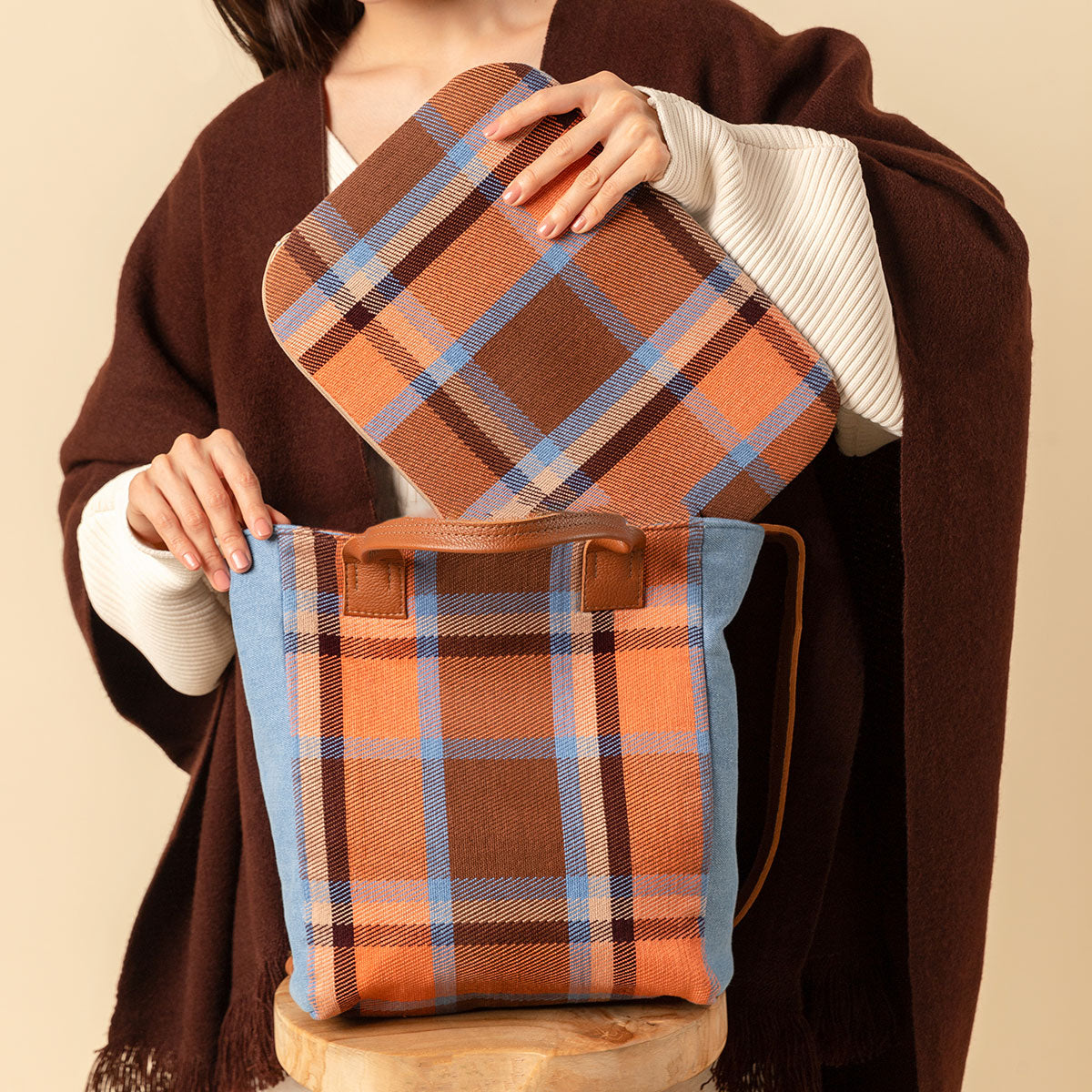 Hand woven artisan Ana Tote and Elenita Laptop Case in Brickstone pattern. A model puts the Elenita Laptop Case into the Ana Tote. The brickstone pattern is a plaid with brown, orange, and sky blue colors. The model is wearing a brown shawl over a white sweater.