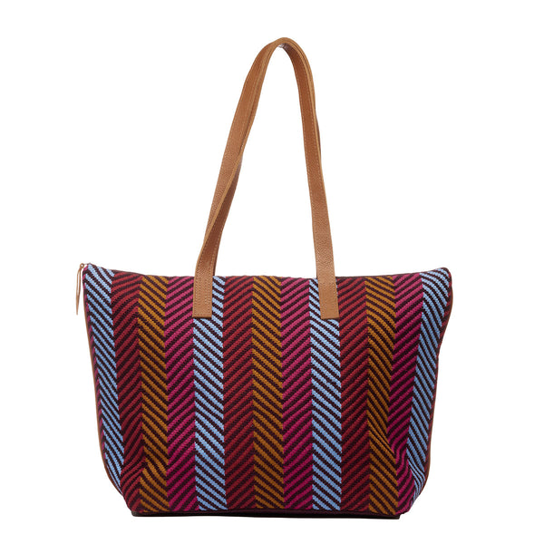 Hand woven artisan Estella Tote in Winter Berry pattern. The front is shown. The bag has vertical stripes in blue, red, and ochre yellow colors. It has leather handles and a small leather zip pull to the right.