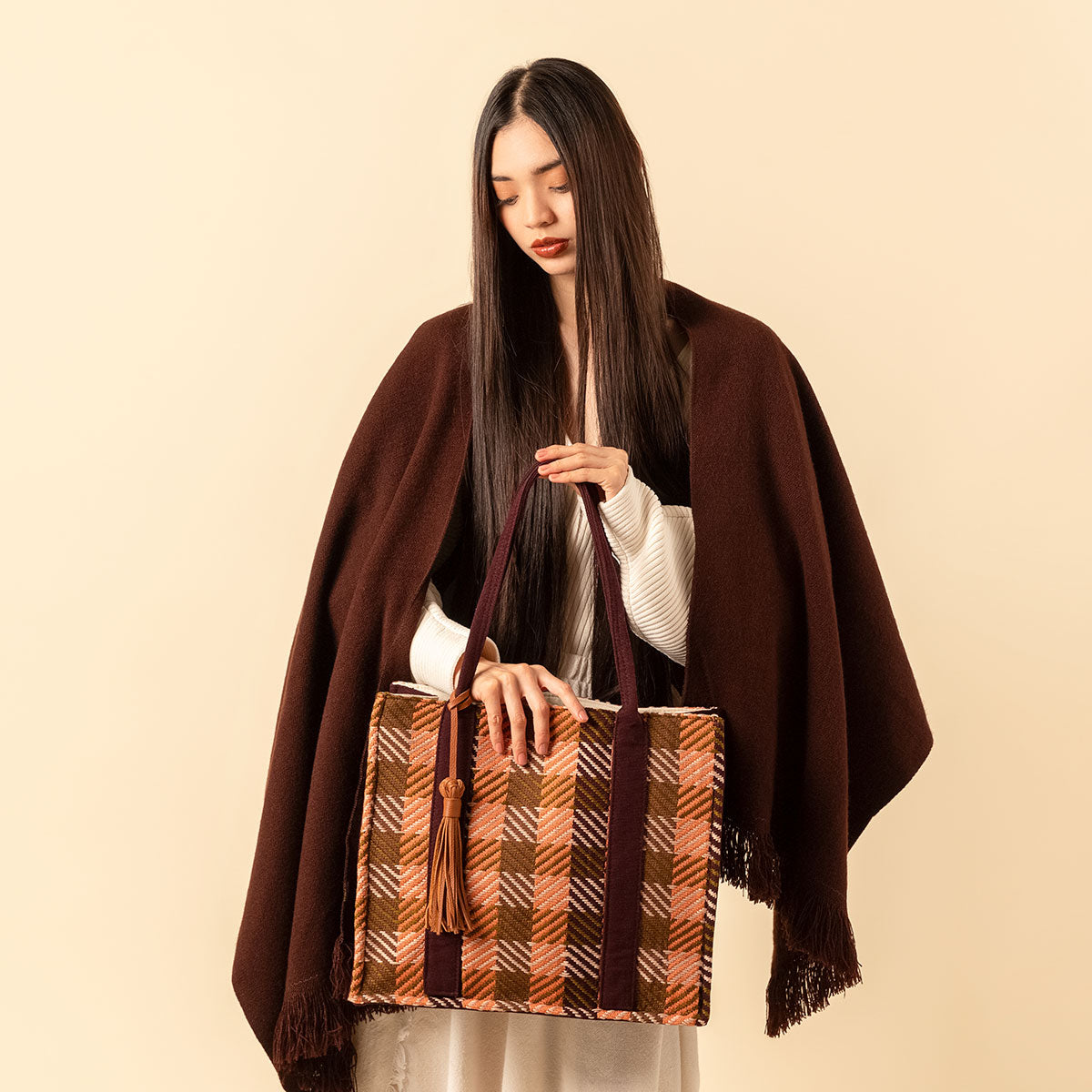 Hand woven artisan Irma Tote in Moss Weave pattern. A model holds the tote bag and looks down with a shawl drapped over her. The moss weave has an orange and olive green plaid. It has a leather tassel attached to the dark brown handle.