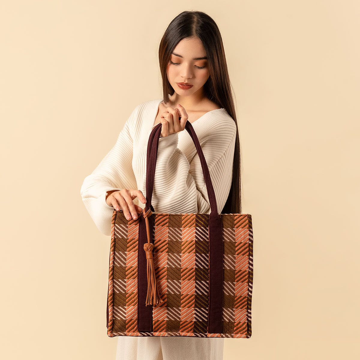 Hand woven artisan Irma Tote in Moss Weave pattern. A model holds the tote and looks down. The moss weave pattern is an orange and olive green plaid. A leather tassel is attached to the dark brown handles.