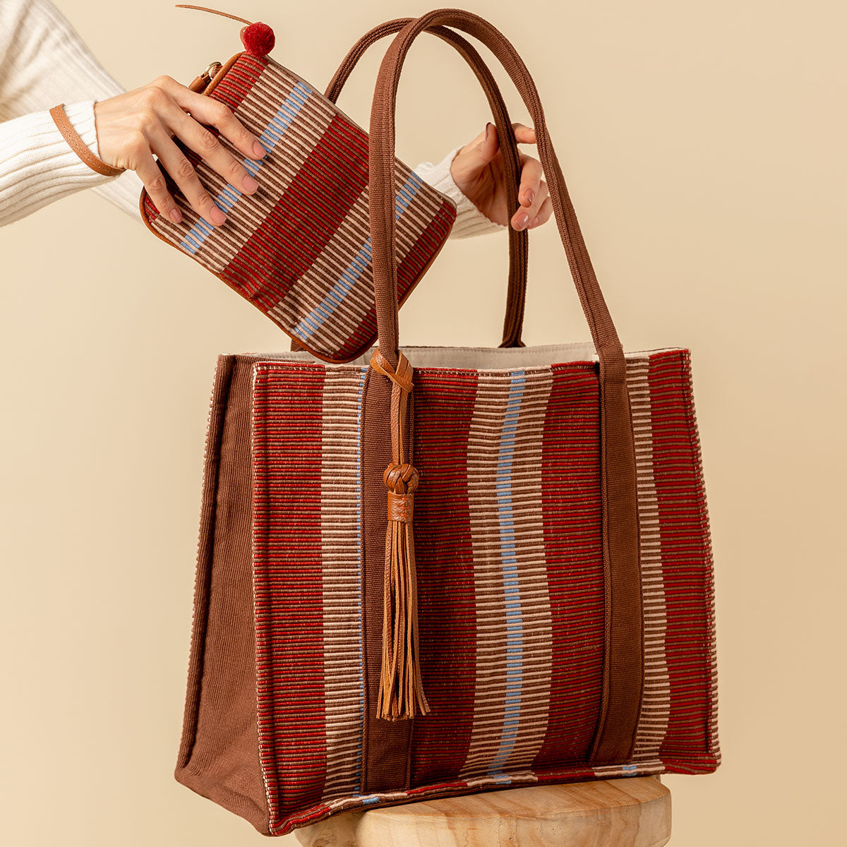 Hand woven artisan Irma Tote and Mini Lily Wrislet in Chestnut color. A model is shown putting the wristlet into a tote. The Chestnut pattern has vertical burgundy, tan, and beige stripes. It has brown solid color sides and handles. 