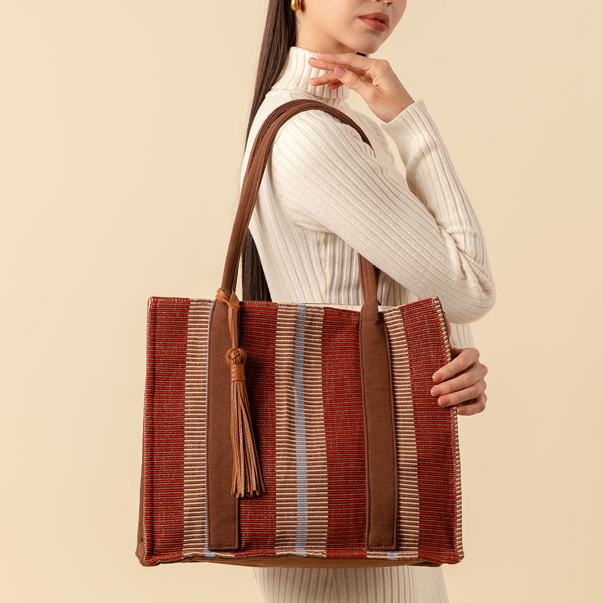 Hand woven artisan Irma Tote in Chestnut color. A model carries the tote over her shoulder. The bag has burgundy, beige, and tan vertical stripes. It has a leather tassel attached to the brown handles.