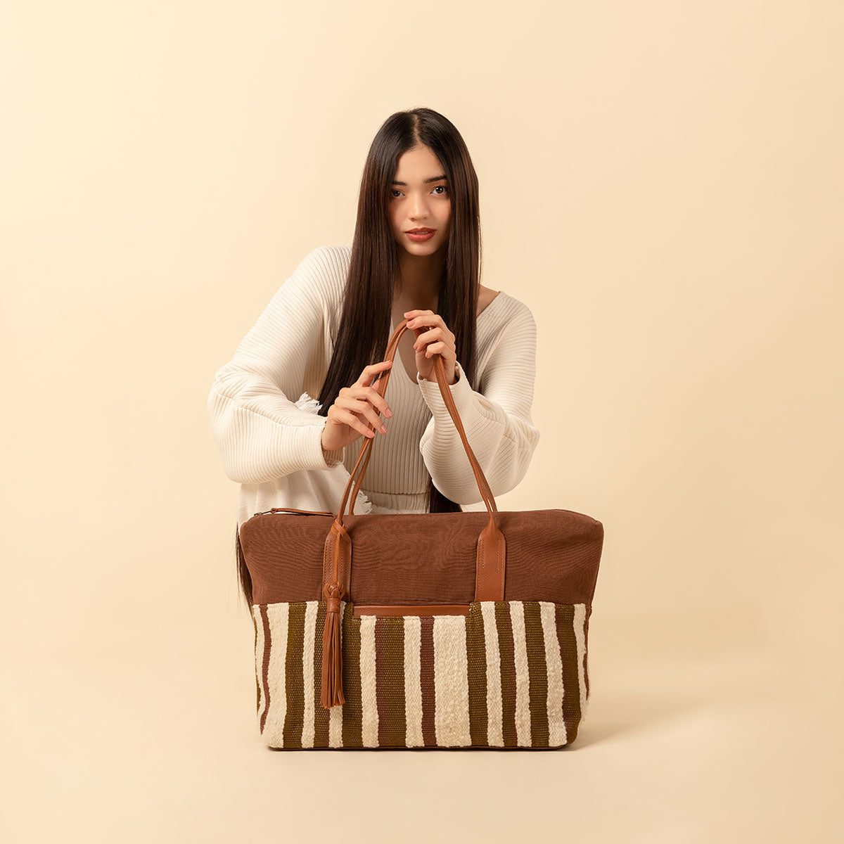 Hand woven artisan Soledad Weekender in Forest pattern. A model crouches down and holds the Soledad Weekender bag, looking into the camera. The bag has vertical beige and olive green stripes with a horizontal brown fabric. It has leather handles, a tassel, and a leather zip pull.