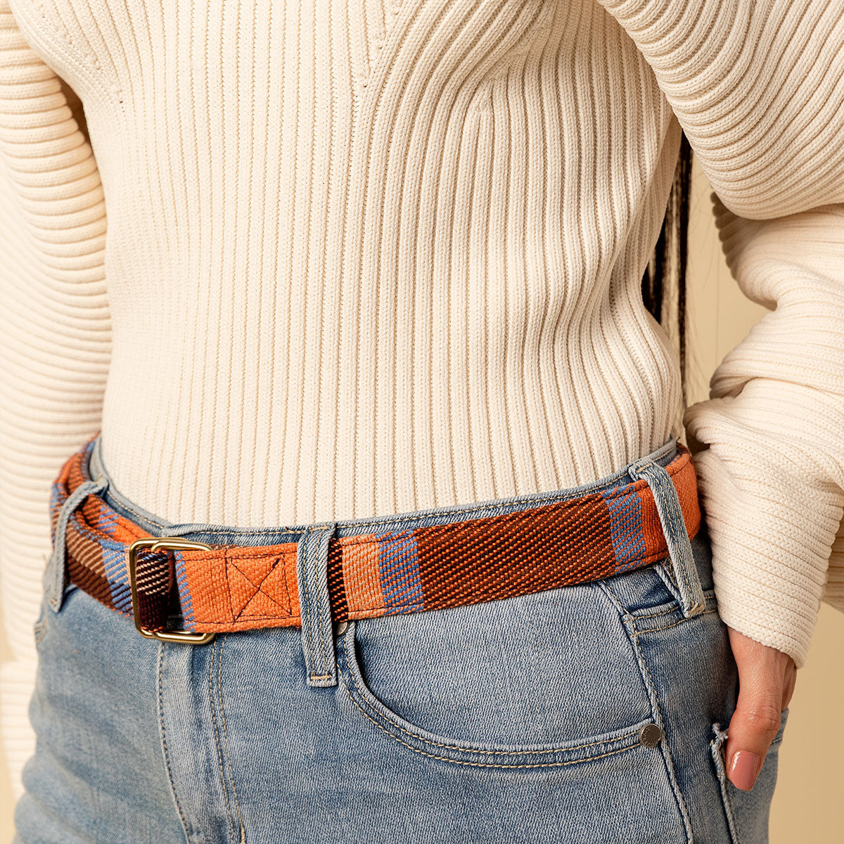 Hand woven artisan Celeste belt in Brickstone color. A model wears the orange and brown plaid belt. The photo is cropped to show the belt holding up jeans and a shirt.