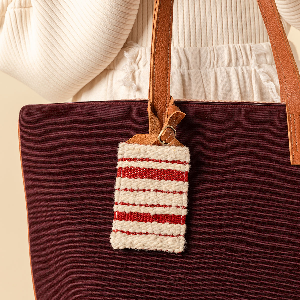 Hand woven artisan Ines Luggage Tag in Cherry Red pattern. The tag has a white and red horizontal stripped pattern with a leather loop attached to a tote bag. A model holds the tote with the tag. The model wears a light beige sweater and pants.