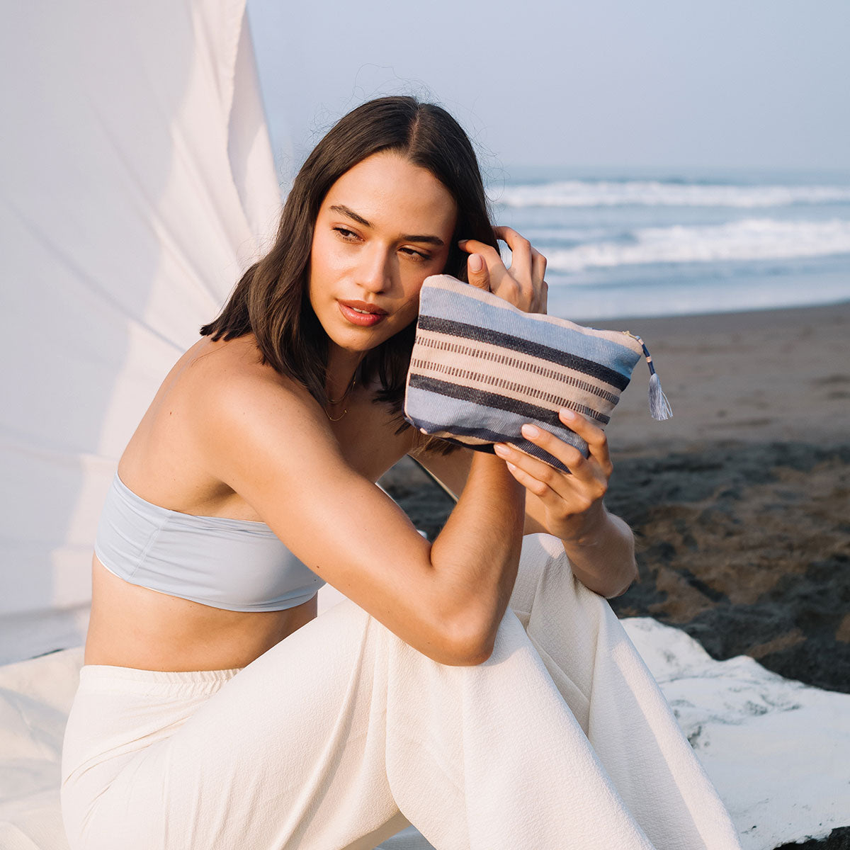 A model sits and holds the Mini Cristina Pouch in Skyscape. The background is a white tent on the beach.