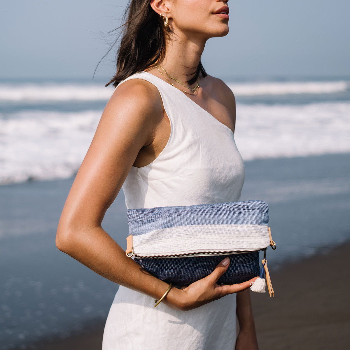 A model holds the folded Florentina Clutch-to Hobo (Ocean Breeze) and stands sideways, profile view. She is on the beach.