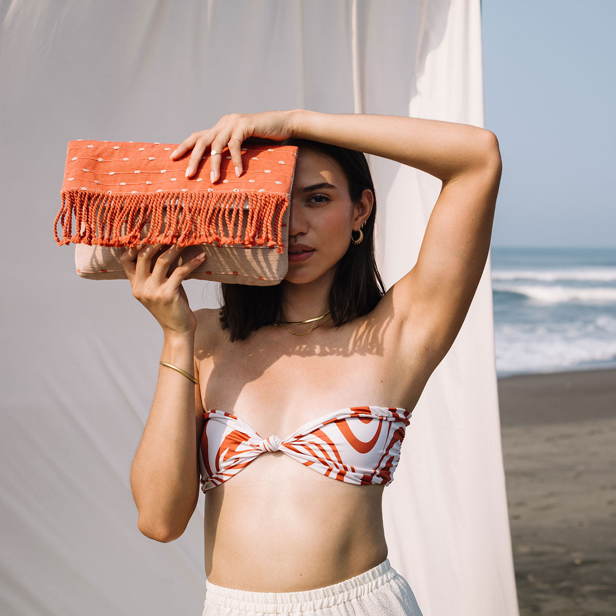 A model holds the Margarita Clutch (Desert Sands) over one eye. She stands in front of a white tent on the beach.