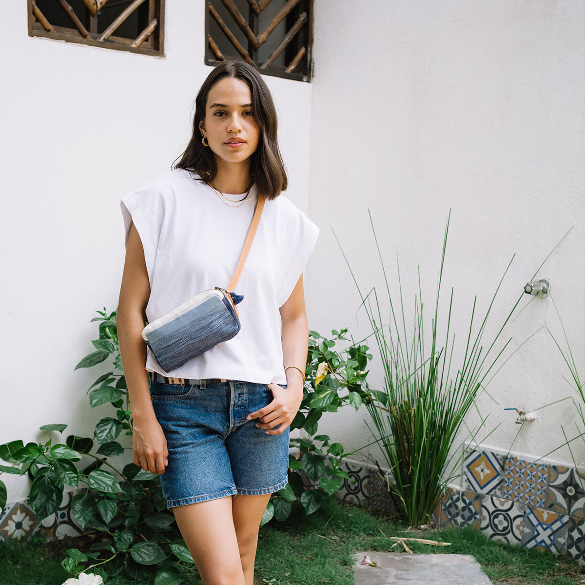 A model stands and wears the Felipa Belt Bag in Ocean Breeze as a crossbody around the torso. The background is a white-walled courtyard with plants and wooden windows.