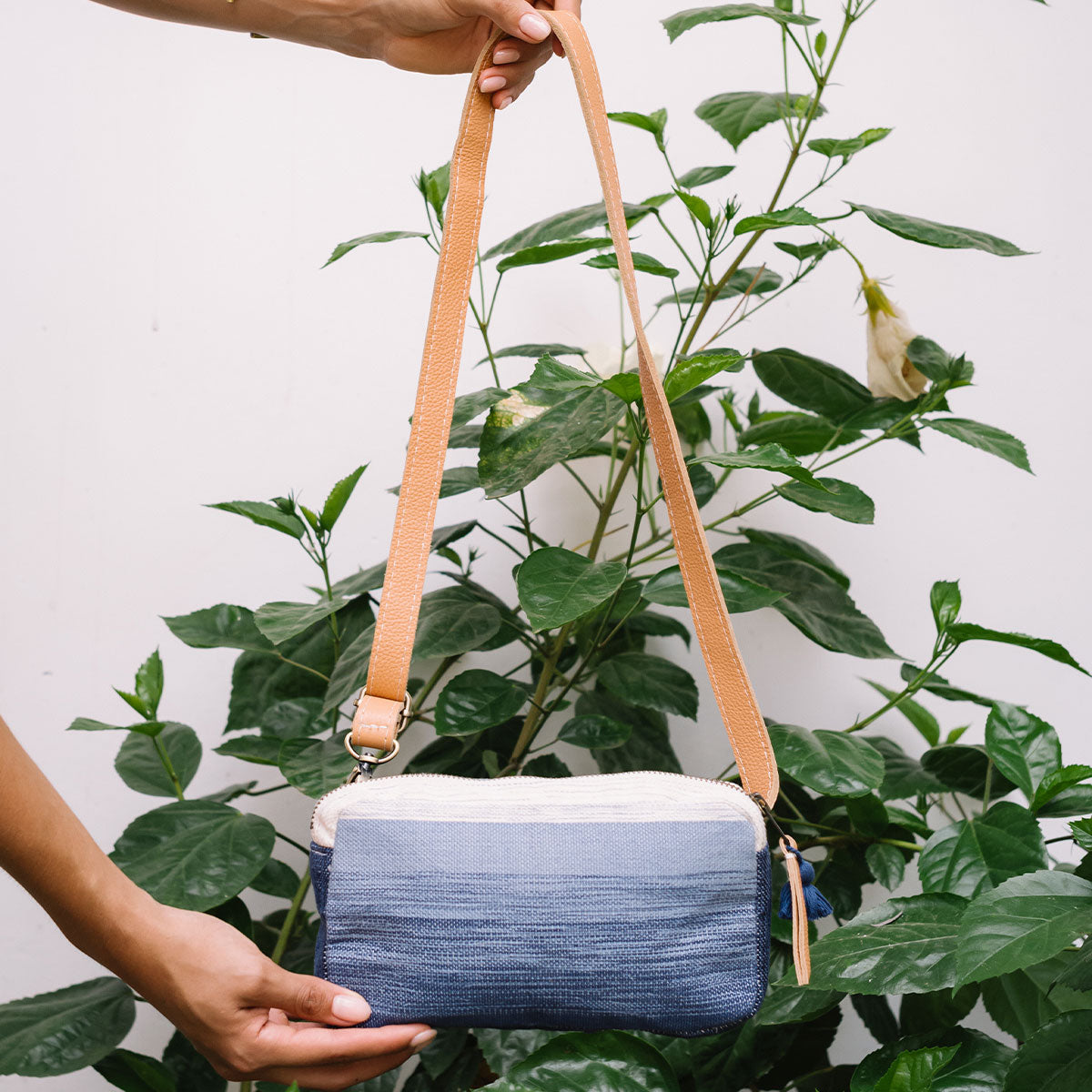 A model holds the Felipa Belt Bag (Ocean Breeze) by the top strap, showing the full length of the leather strap. The background is a white wall with plants.