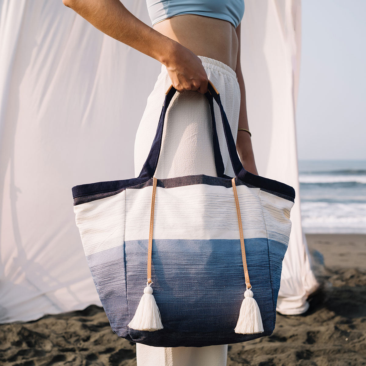 A model stands sideways and holds the Rosa Tote in Ocean Breeze. The background is a white tent in front of the seashore.