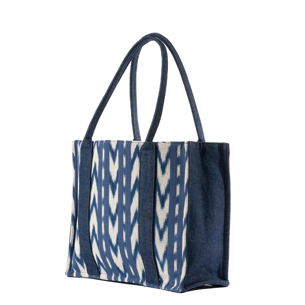 A right-side angle of the Irma Tote in Atitlán Hills pattern. It has vertical and chevron dark blue and white stripes. It has a rectangular shape with a flat bottom. The sides and handles are a solid dark blue color.