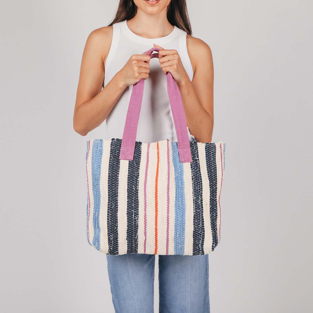 A model holds the hand woven artisan Patricia Interwoven Tote in Spring Sherbert. It has a sky blue, dark blue, pink, and orange vertical striped pattern. It has pink handles.